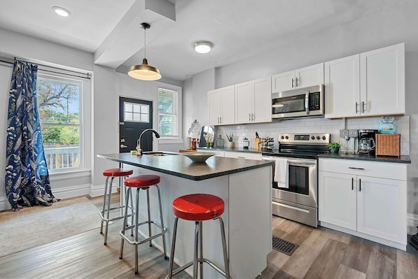 A modern kitchen with white cabinets, a central island with stools, stainless steel appliances, and a large window with blinds.
