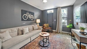 Una sala de estar moderna con un sofá, una mesita redonda, un ventilador de techo, una lámpara, una silla, una ventana con cortinas y un cuadro de bicicleta colgada en la pared.