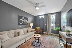 Ein modernes Wohnzimmer mit einer Couch, einem runden Couchtisch, einem Deckenventilator, einer Lampe, einem Sessel, einem Fenster mit Vorhängen und einem Fahrradbild an der Wand.