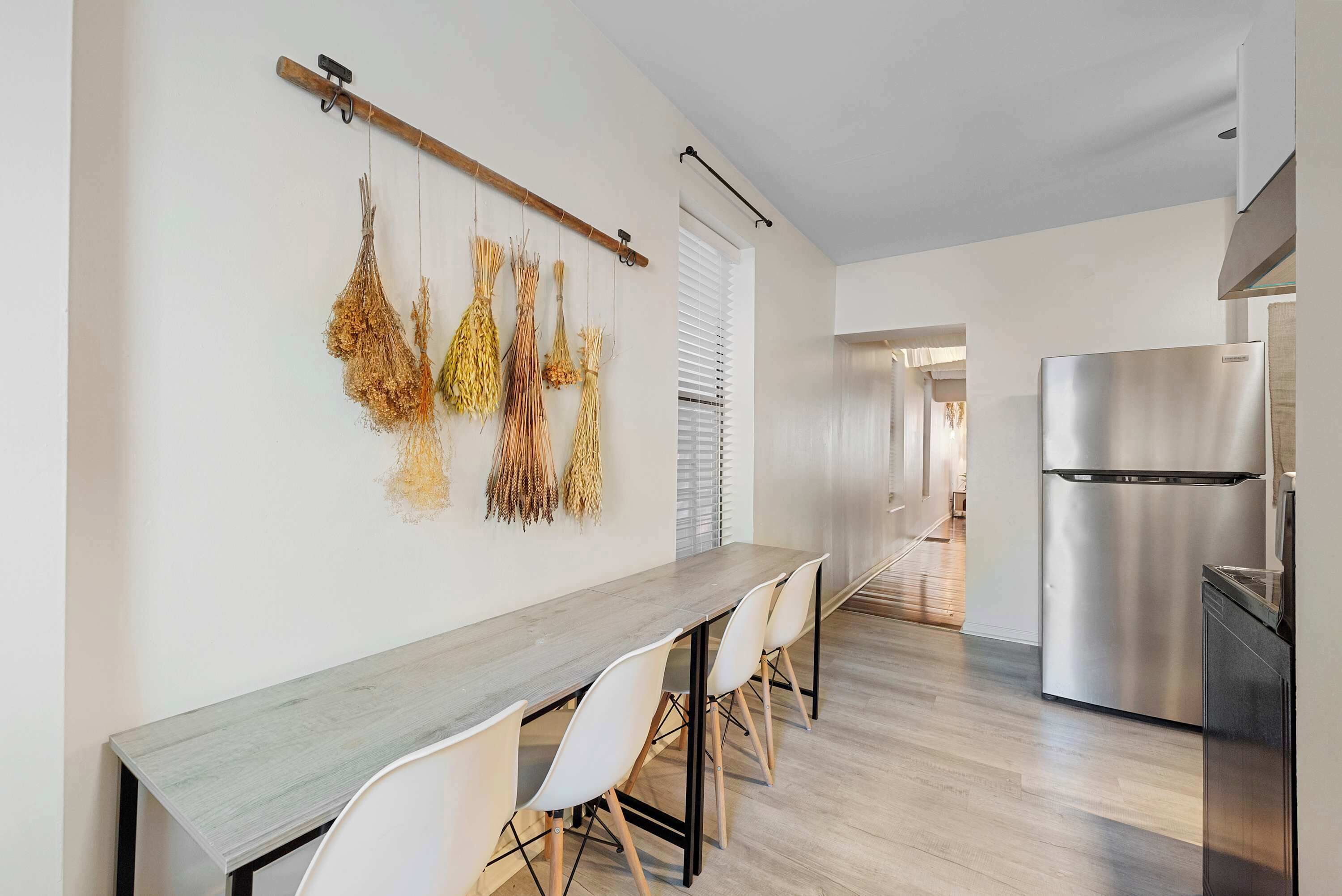 A modern kitchen with a stainless steel refrigerator, a wooden dining table with white chairs, and dried plants hanging on the wall.