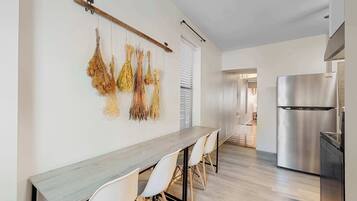 A modern kitchen with a stainless steel refrigerator, a wooden dining table with white chairs, and dried plants hanging on the wall.