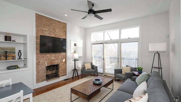 Una sala de estar moderna con chimenea de ladrillo, televisor de pantalla plana, ventilador de techo y ventanas amplias.