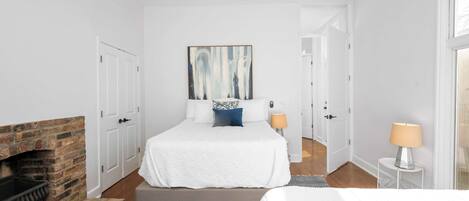 Une chambre avec une cheminée en briques, un lit avec une literie blanche, un oreiller bleu et un tableau accroché au mur.
