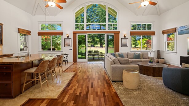 A spacious living area with a large window, a kitchen with a bar, and a ceiling fan. - Lei Aloha - A Private Retreat, Pool + Fairway views (Princeville)