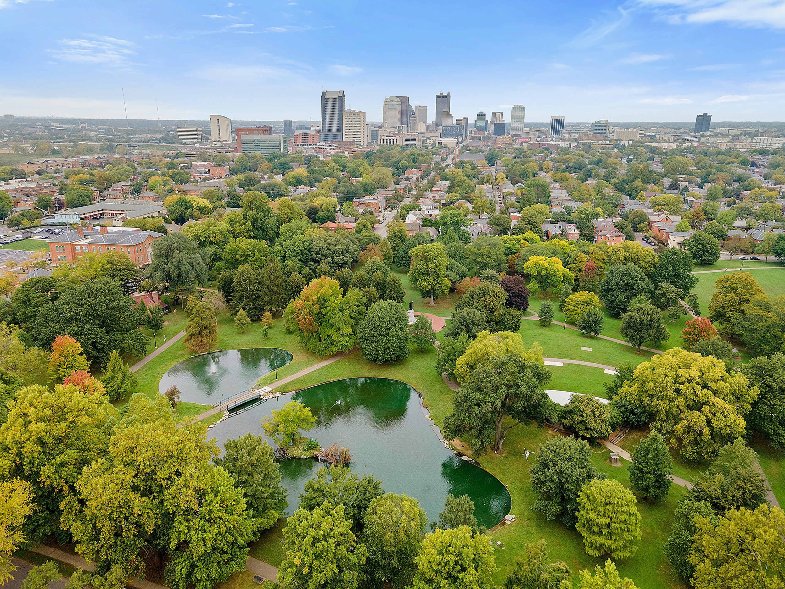 Un parco con uno stagno, circondato da alberi e uno skyline cittadino sullo sfondo.