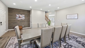 A dining area with a long table, multiple chairs, a wooden accent wall, a framed quote, and a staircase in the background. - Ark & Creation Retreat – For 12! (Walton)