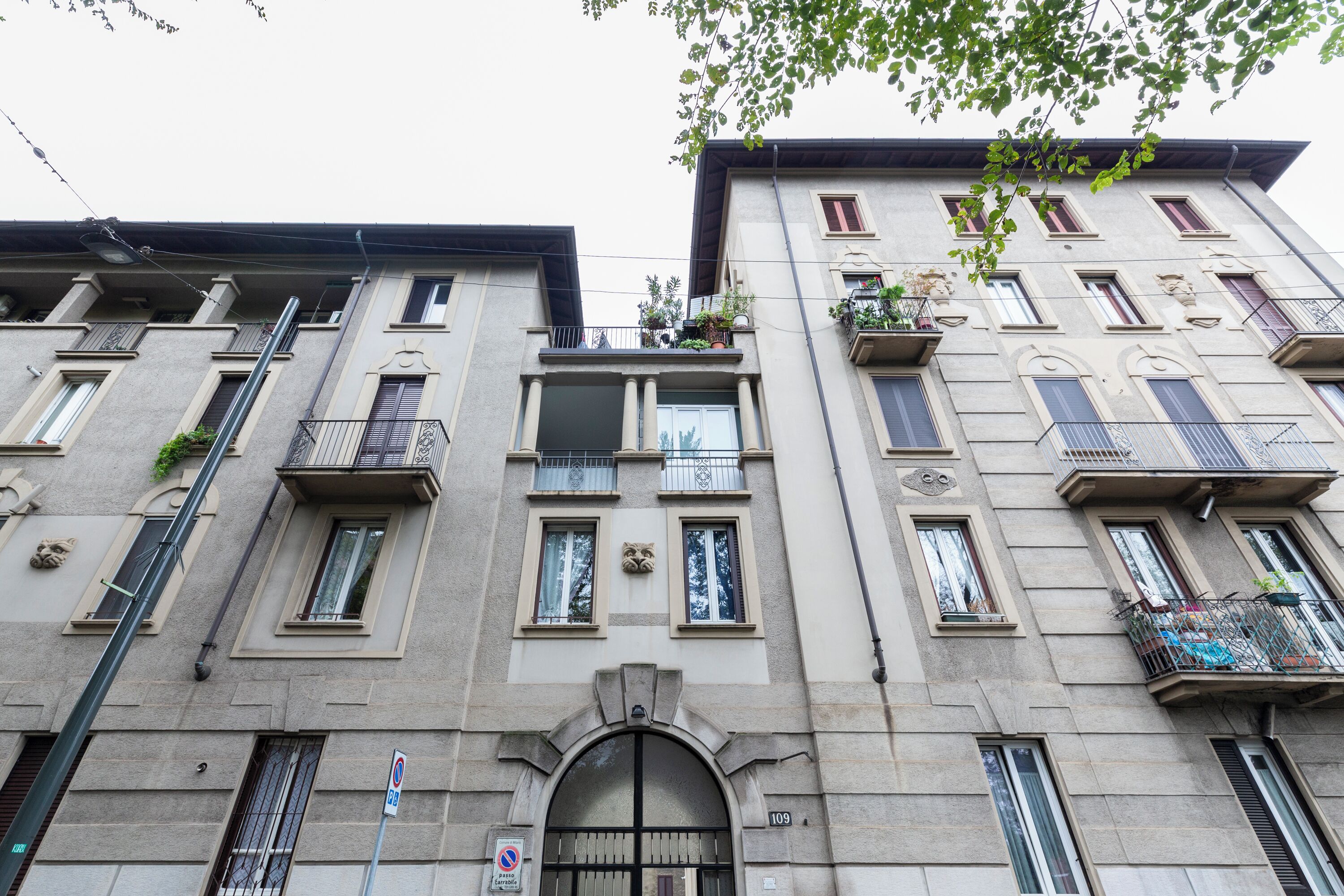 A multi-story building with a classic architectural style, featuring balconies with plants, a central entrance with an archway, and a visible address number.