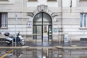 Une entrée d’immeuble avec un scooter garé, un panneau indiquant l’interdiction de se garer et un reflet sur le sol mouillé.