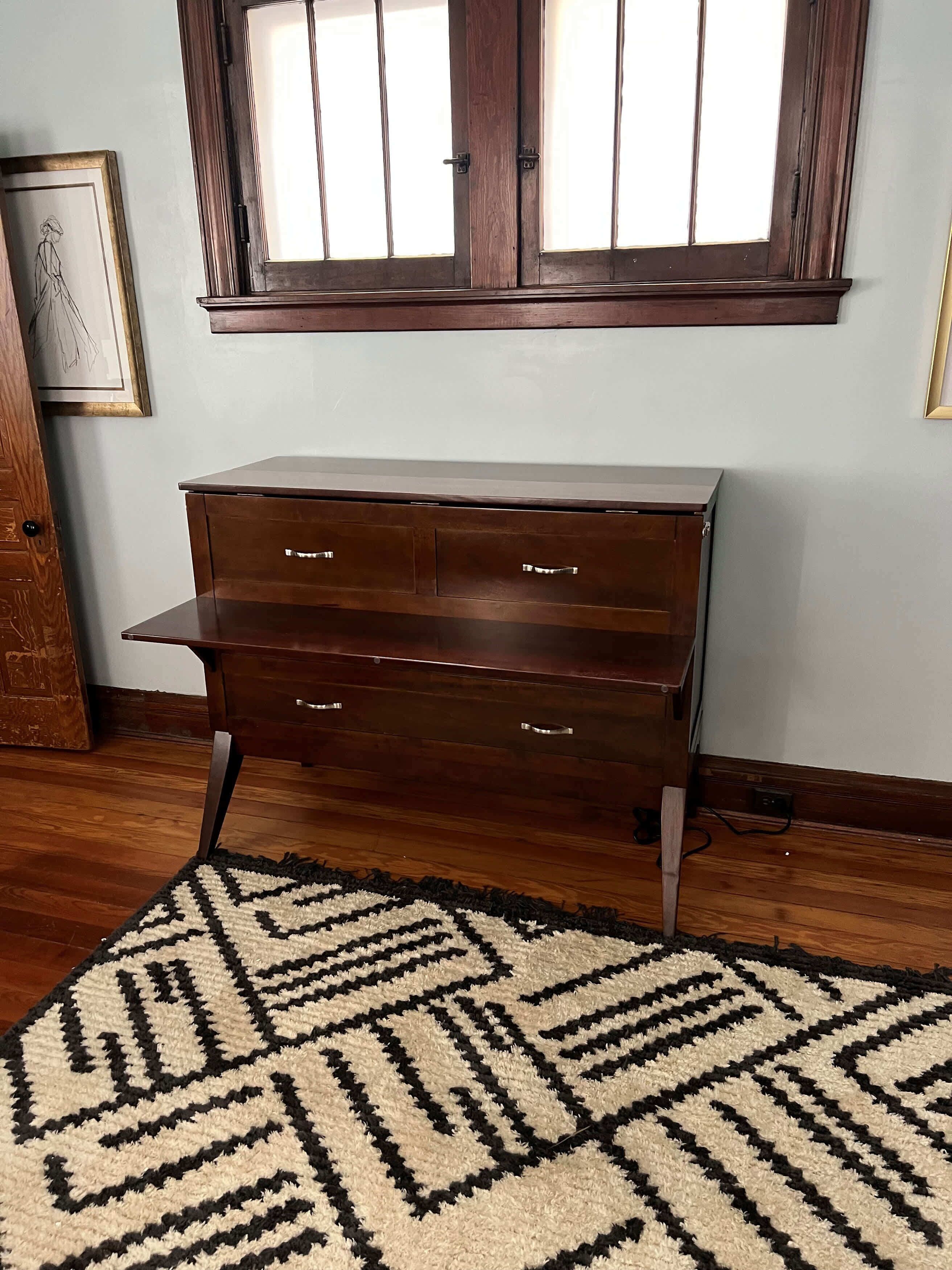 A wooden dresser with drawers against a wall, a window above, and a patterned rug on the floor.