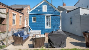 Ein blaues Haus mit weißer Tür und Fenstern, einem überdachten Grill und einem Korbstuhl.