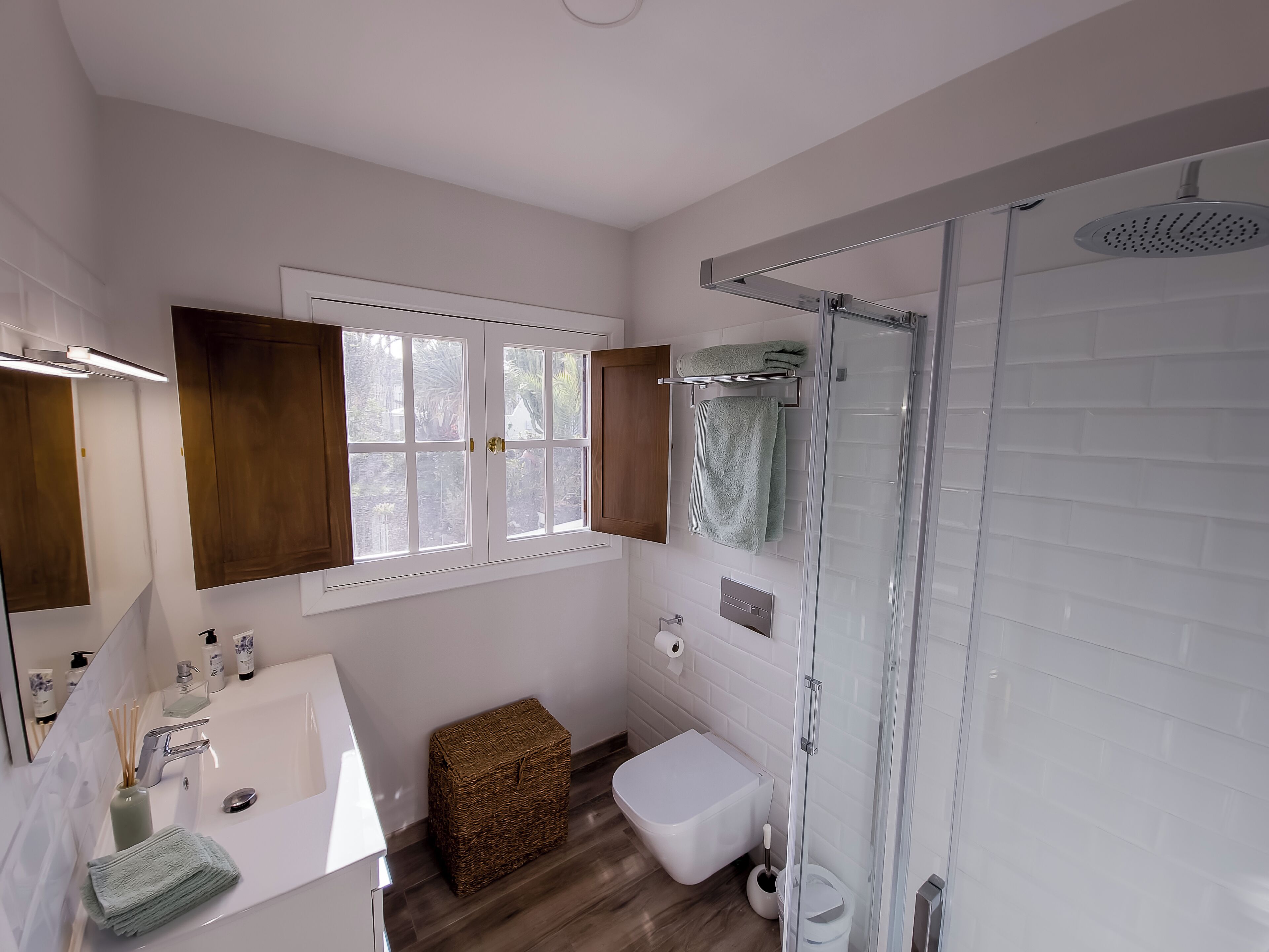 Ein modernes Badezimmer mit Waschbecken in Weiß, einer Duschkabine aus Glas, einer Toilette und einem Fenster mit Blick auf Grünflächen.