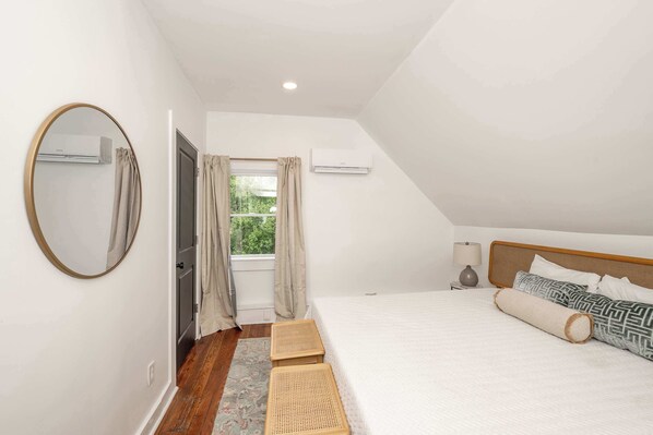 A bedroom with a large bed, a round mirror, a window with curtains, and an air conditioning unit.