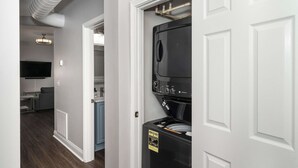 A laundry room with a front-loading washer and dryer, a white door, and a hallway leading to a living area with a flat-screen TV. - Heart of Mainstrasse 3 (Covington)