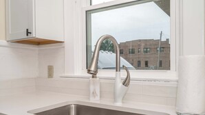 A kitchen with a stainless steel sink, a white countertop, a window with a view of buildings, and a paper towel dispenser. - The Mariemont Cottage (Cincinnati)