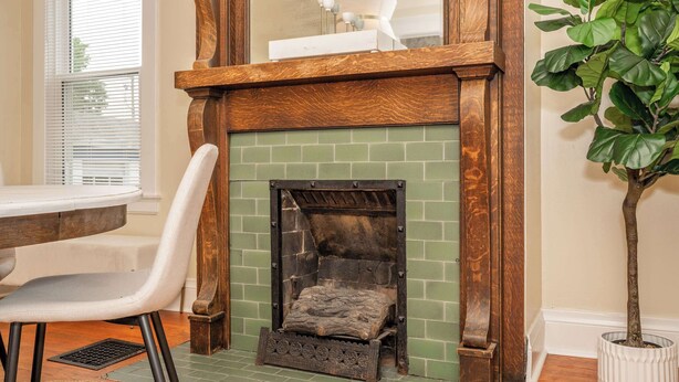 A fireplace with a green brick surround, a wooden mantel, and a white chair in front. - The Mariemont Cottage (Cincinnati)