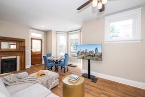 A living room with a fireplace, a dining area with blue chairs, a television mounted on the wall, and a sofa with a hat and sunglasses on it. - Near Cincy | Skyline Views | Historic 4BR (Ludlow)