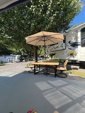 A covered picnic table with chairs and a hanging flower basket, set up under a tree in a campground with RVs in the background. - Cozy 2-bedroom RV with WiFi, AC in charming Gatlinburg (Gatlinburg)