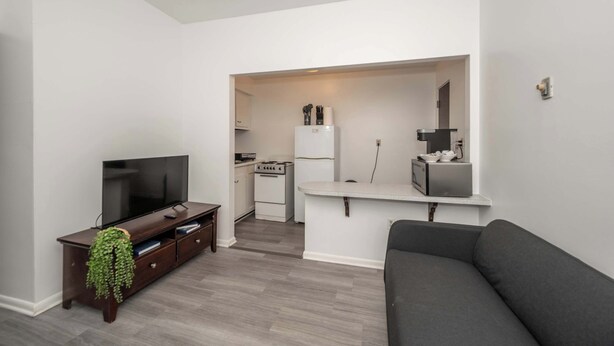 Een moderne woonkamer met een flatscreen televisie, een donker houten entertainmentkast en een grijze bank. In de achtergrond is een keuken te zien met witte apparatuur en een klein eiland.