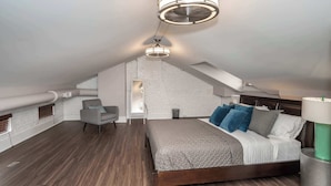 Un dormitorio estilo loft moderno con techo inclinado, paredes de ladrillo visto, suelo de madera, una cama con muchos almohadones y un ventilador de techo.