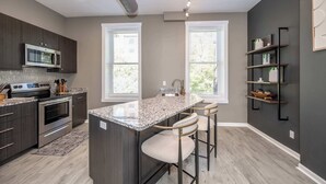 A modern kitchen with dark cabinetry, a built-in microwave, a stainless steel oven, and a granite countertop island with bar stools. - Queen City Views Suites (Cincinnati)