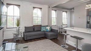 Ein moderner Wohnraum mit grauem Ecksofa, einem Glastisch, einer Bar mit Hockern und großen Fenstern mit Jalousien.