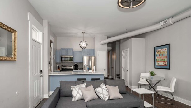 Ein modernes Wohnzimmer mit einem grauen Ecksofa, einem kleinen weißen Tisch und einem Glastisch. Im Hintergrund ist eine Küche mit blauen Schränken und Edelstahlgeräten zu sehen.