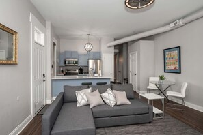 A modern living room with a grey sectional sofa, a small white table, and a glass coffee table. A kitchen with blue cabinets and stainless steel appliances is visible in the background. - Heart of Mainstrasse Suites (Covington)
