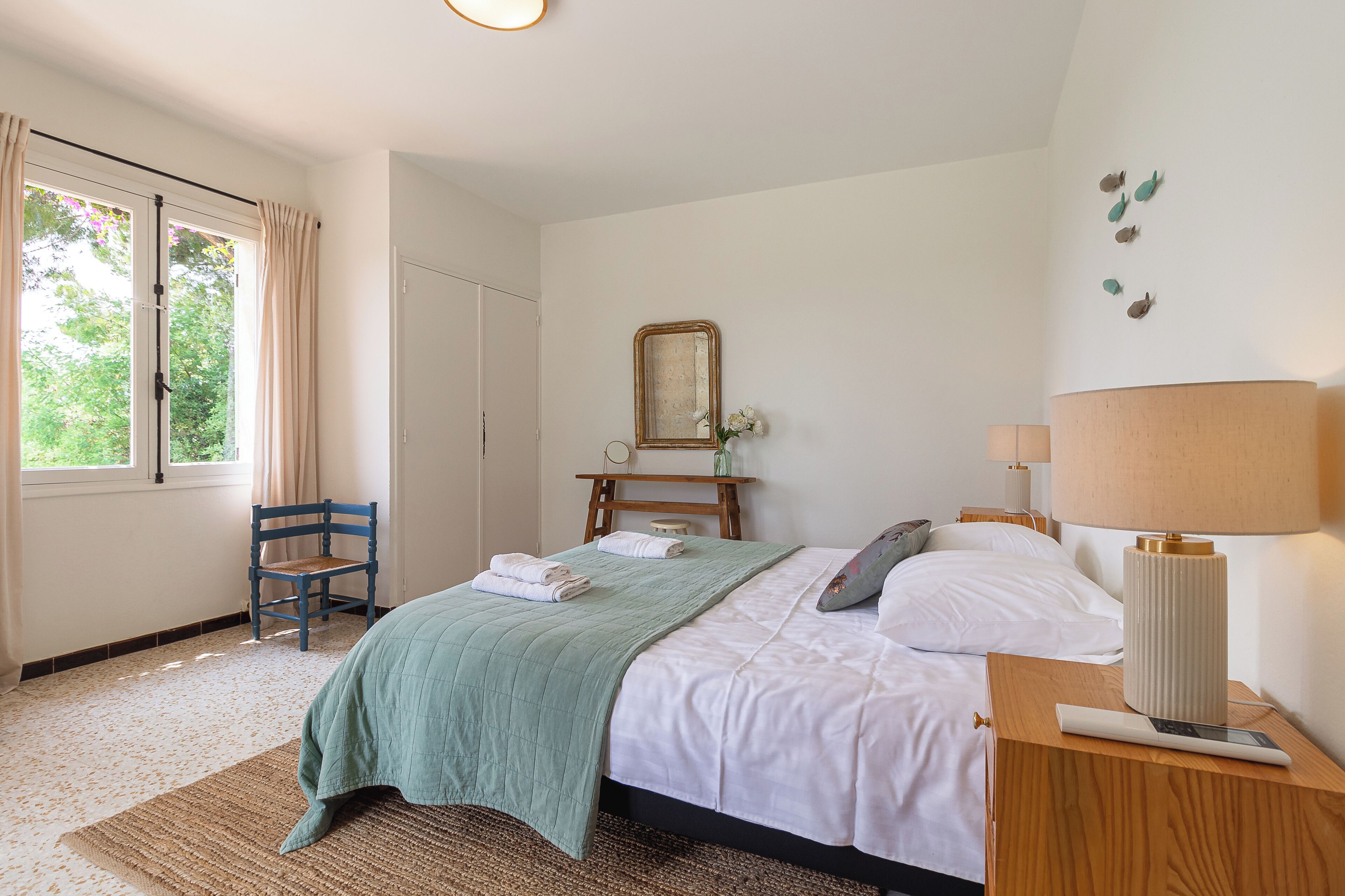 A neatly arranged bedroom with a bed, a chair, a bedside table with a lamp, and a window with curtains.