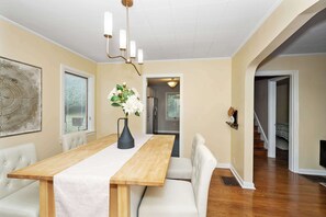 A dining area with a wooden table, white upholstered chairs, a vase with flowers, and a chandelier.