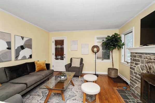 A living room with a sofa, coffee table, and two white stools. There is a fireplace, a large plant, and a floor lamp.