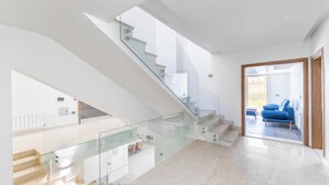 Un intérieur moderne avec un escalier en verre menant à un deuxième étage, un canapé bleu dans l’espace de vie et une vue à travers une porte ouverte.