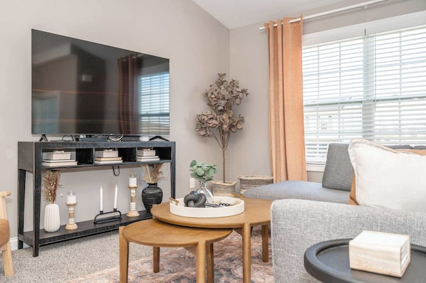 A modern living room with a flat-screen TV, a dark wooden console table, a grey sofa, and a large window with blinds.