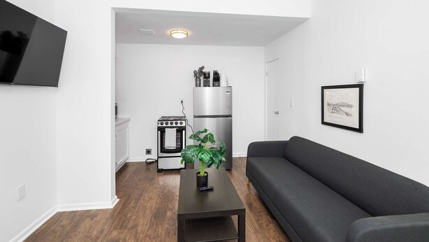 A modern living room with a grey sofa, a small table with a plant, a kitchen area with a stove, oven, and refrigerator, and a mounted TV. - Northside Suites 4 – Your Perfect Vacation Haven! (Cincinnati)