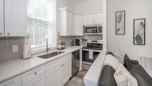 A modern kitchen with white cabinets, a built-in microwave, and an oven. There is a dining area with a gray sofa and a small table. - Covington Charm 201 (Covington)