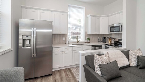 A modern kitchen with white cabinetry, stainless steel appliances, and a grey sofa with patterned cushions. - Covington Charm 201 (Covington)