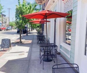 A row of outdoor tables and chairs under red umbrellas, with a sign reading "TASTE" above.