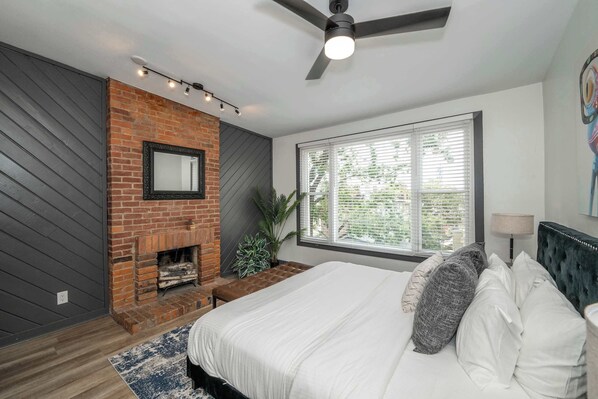 Un dormitorio con chimenea de ladrillo, una cama grande con ropa de cama blanca, un ventilador de techo y una ventana con persianas.