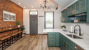 A kitchen with green cabinets, a marble countertop, and a brick wall. - 2 King Beds | Near OTR + Downtown | Free Parking (Cincinnati)