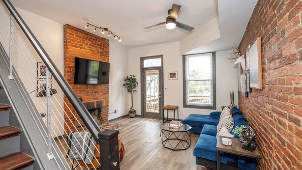 A modern living room with a brick fireplace, a blue sofa, a glass coffee table, and a staircase with metal railings. - 2 King Beds | Near OTR + Downtown | Free Parking (Cincinnati)