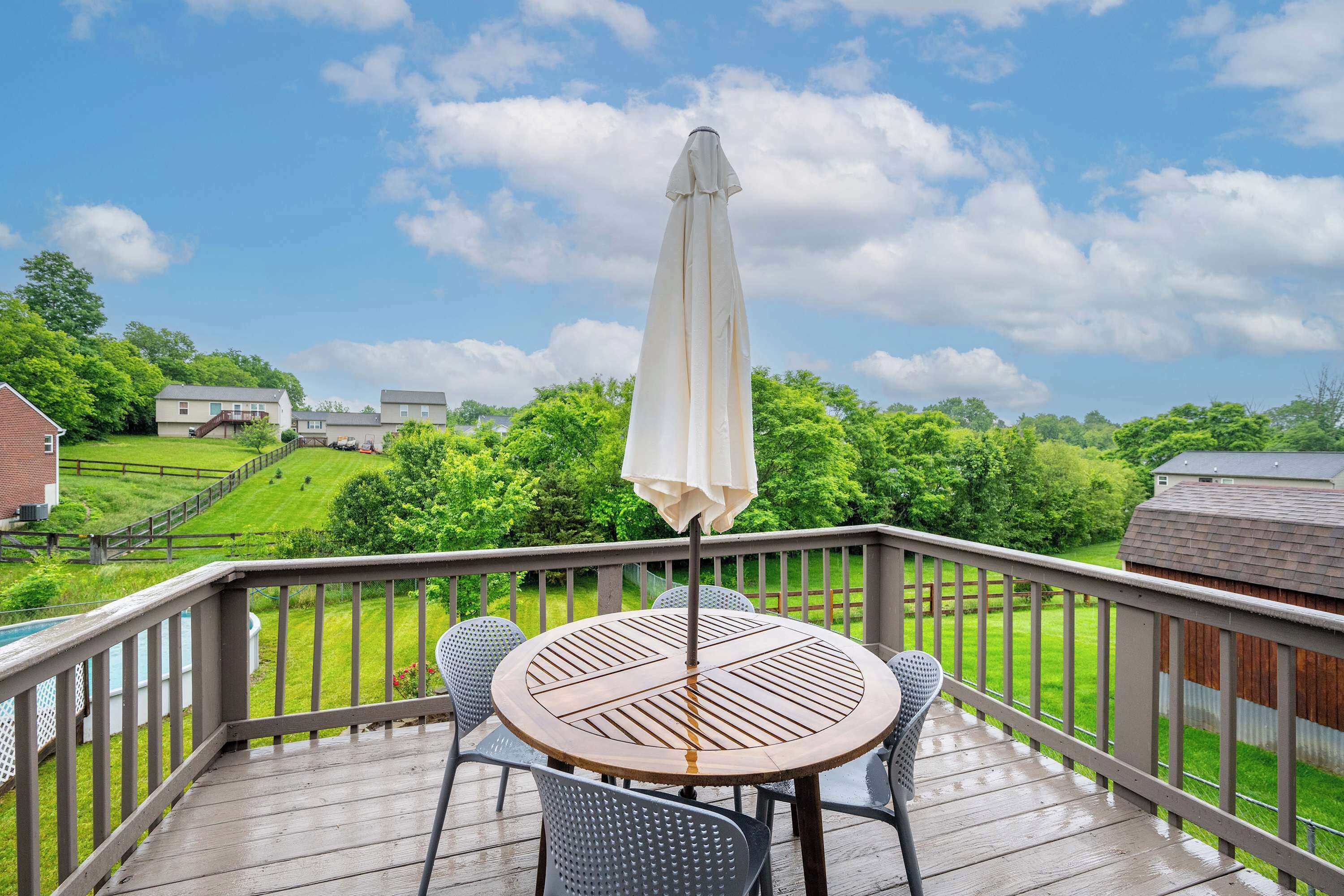 A wooden deck with a round table and chairs, an umbrella, and a view of a green landscape with houses in the background.