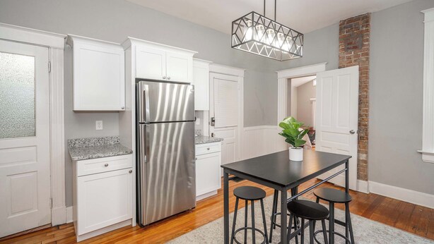 A modern kitchen with white cabinets, a stainless steel refrigerator, a black dining table with stools, and a hanging light fixture. - Suites on Main 1 (Covington)