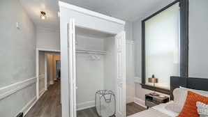 A bedroom with a bed, a bedside table, a lamp, a window with blinds, a closet, and a hallway leading to another room. - Chic Modern Gem w/ Style Suites (Cincinnati)