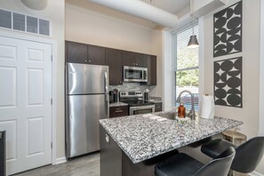 A modern kitchen with a stainless steel refrigerator, microwave, and oven, a granite countertop, and a black pendant light. - OTR 1BR | Private Balcony | Near Downtown (Cincinnati)