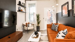 A modern living room with a sofa, coffee table, and a TV. There is a ceiling fan, a window with blinds, and a door.