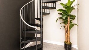 A spiral staircase with a black metal railing, a potted plant with green leaves, and a light-colored wall. - OTR 1BR | Private Balcony | Near Downtown (Cincinnati)