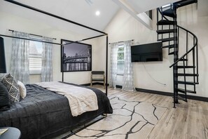 Une chambre à coucher avec un lit, un téléviseur fixé au mur, un escalier et une fenêtre avec des rideaux.