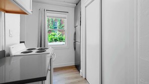 A narrow kitchen with a white stove, a window with a view of greenery, and a refrigerator.