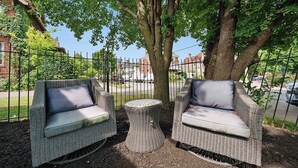Two wicker chairs with cushions, a small round table, and a tree in a fenced outdoor area.