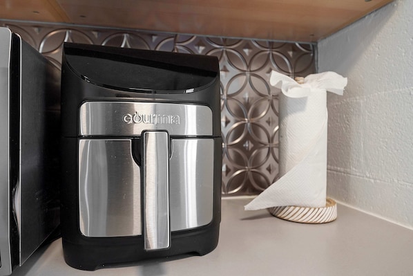 Una cafetera compacta y moderna con acabado metálico, ubicada sobre una encimera junto a un salpicadero de azulejos decorativos y un dispensador de pañuelos.