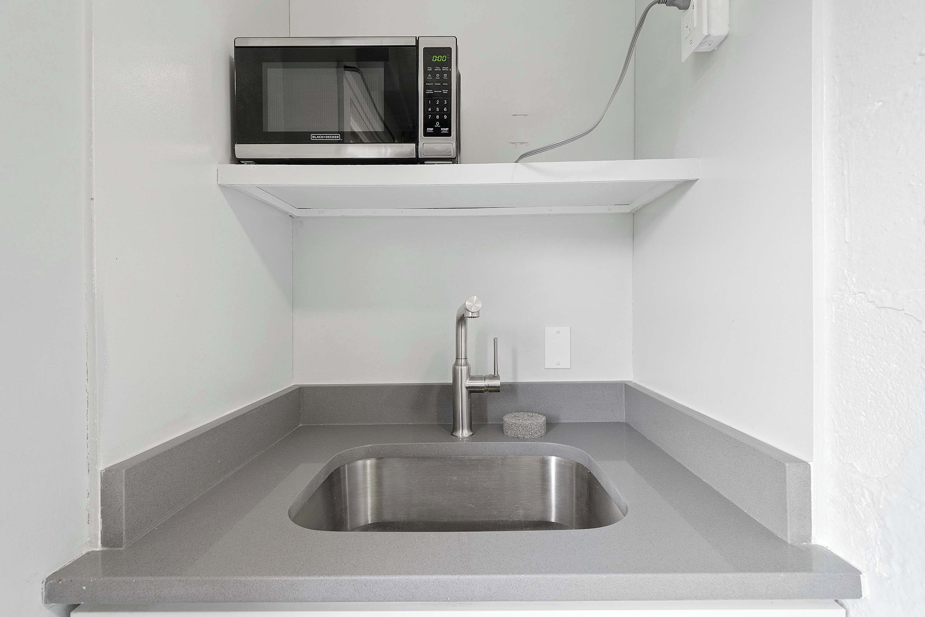 A compact kitchen with a built-in microwave, a stainless steel sink, and a single electrical outlet on the wall.